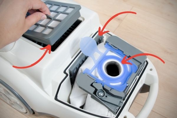 Öppnad dammsugare med pilar som visar vad man bör kontrollera. Dammsugarpåse, mikrofilter och hepafilter